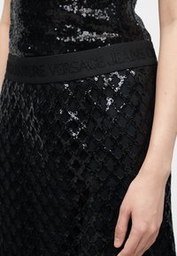 Μαύρο τοπ με παγιέτες, συνοδευόμενο από μαύρη δίχτυνη φούστα και φαρδύ ζωνάρι με ανάγλυφη επιγραφή "Versace Jeans Couture", το χέρι ακουμπάει στο πλάι.