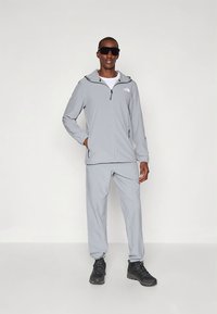 Šviesiai pilkas vėjo nepraleidžiantis striukė su užtrauktuku priekyje, gobtuvu ir šoninėmis kišenėmis, derinamas su atitinkamomis pilkomis kelnėmis ir juodais žygių batais.