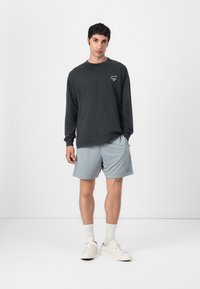 Donkere grijze shirt met lange mouwen en een grafische print, gecombineerd met lichtgrijze shorts. Witte sneakers en kniehoogte witte sokken maken de look compleet.