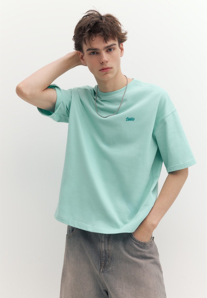 Jeune homme aux cheveux courts et bruns portant un t-shirt oversize bleu clair, un collier chaîne argenté et un jean gris, posant avec une main derrière la tête.