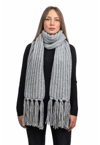Grå stickad scarf med en tjock textur, med långa fransar i varje ände. Bärs virad runt halsen. Materialet ser mjukt och varmt ut.