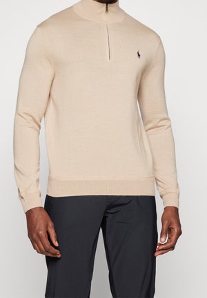 Pull beige à manches longues avec fermeture éclair quart de zip, petit logo bleu marine sur la poitrine gauche, porté avec un pantalon noir.