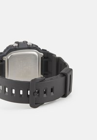 Montre en caoutchouc noir avec un boîtier rectangulaire en métal argenté, bracelet texturé avec découpes et fermoir robuste.