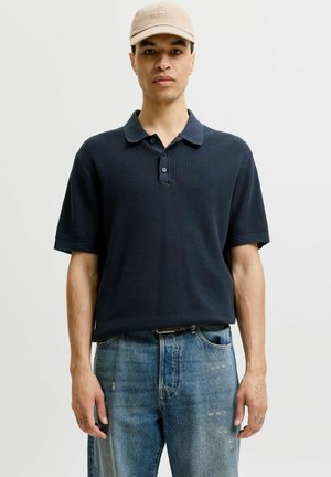 Jeune homme portant une casquette beige, un polo à manches courtes texturé foncé et un jean bleu clair usé, debout devant un fond uni.