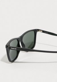 Óculos de sol de plástico preto com lentes verdes. Armações quadradas com hardware mínimo. Apresenta a marca "Polaroid" na lateral.