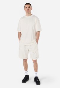 Lyscreme oversized T-shirt med korte ærmer, parret med matchende cargo shorts. Sorte sneakers med hvide sokker, der har logoaccenter.