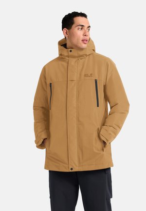 Jonge man die een beige capuchonjack draagt met ritssluitingen op de borstzakken en handen in de zijzakken, kijkt naar rechts tegen een effen achtergrond.