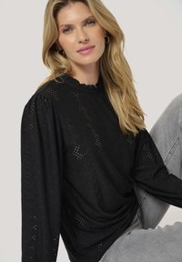 Blusa nera a maniche lunghe con tessuto ad occhio di pernice testurizzato, scollo a volant e maniche a sbuffo. Abbinata a jeans grigi chiari.