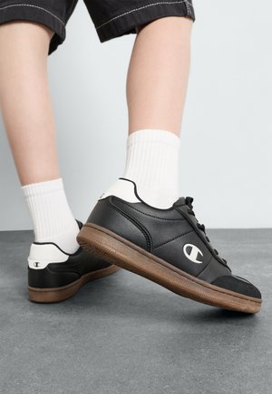 Zapatillas de cuero negro con acentos blancos, con puntera redondeada, diseño de cordones y suela de goma marrón texturizada. Presentan un logo en el lateral.