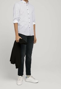 Jasnoniebieska koszula w białe paski z guzikami w kolorze brązowym, z podwiniętymi rękawami; noszona z ciemnymi dżinsami i białymi trampkami, trzymająca czarny sweter.