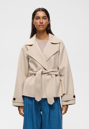 Femme aux longs cheveux foncés portant un manteau en laine beige ceinturé et un jean bleu à jambes larges, debout devant un fond clair uni.