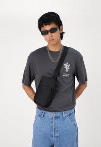 Svart skulderveske med glidelåslomme, båret over en grå T-skjorte med et hvitt blomsterdesign og kombinert med lyseblå denimshorts.