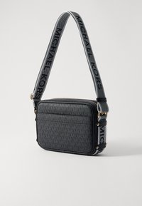 Mustat nahkainen olkalaukku, jossa on geometrinen kuvio. Varustettu kultaisella vetoketjulla ja säädettävällä hihnalla, jossa on "MICHAEL KORS" -teksti.