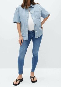 Camisa de mezclilla azul claro, de corte oversize y manga corta, combinada con jeans ajustados azules y chanclas negras. Presenta un bolsillo frontal y botones.