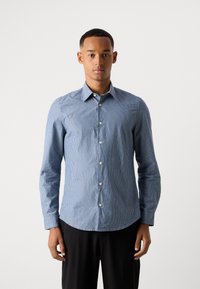 Bărbat îmbrăcat cu o cămașă bleu slim-fit, cu mâneci lungi și dungi albe verticale, purtând pantaloni negri, stand într-un fundal simplu deschis.