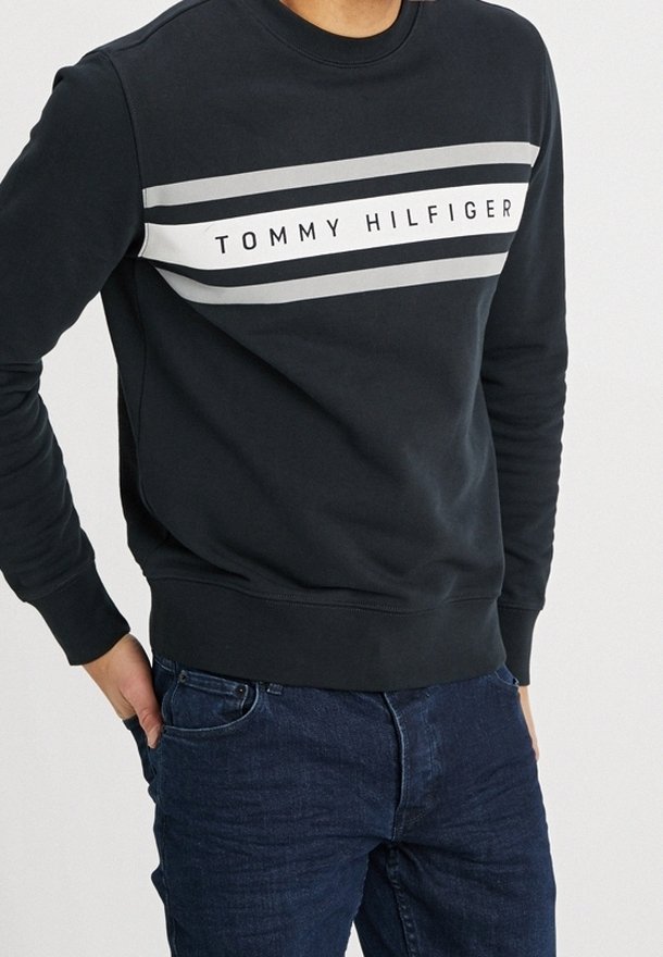Homme portant un sweat-shirt noir avec des rayures blanches et grises et le texte "Tommy Hilfiger", associé à un jean bleu foncé.