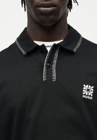 Κοντινό πλάνο ατόμου που φοράει μαύρο πόλο με λευκή ραφή στο γιακά και στο καβαλέτο και λευκό λουλουδάτο λογότυπο HUGΟ στο στήθος.