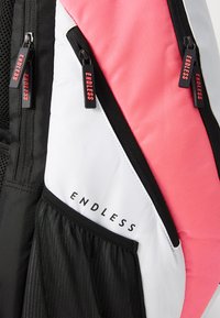 Close-up de uma mochila mostrando um bolso de malha preta, painéis brancos e rosas, e puxadores de zíper pretos com a inscrição "ENDLESS" em texto vermelho.