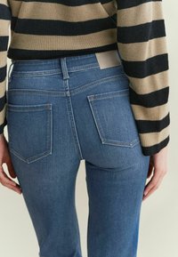 Modre jeans hlače iz denima s srednjim pasom, ozkim krojem in kontrastnim šivanjem. Imajo dve zadnji žepi in rjav usnjen oznako na pasu.