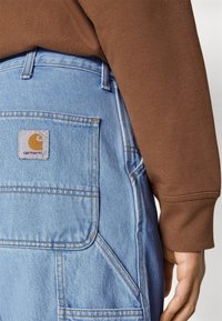 Helle blaue Jeanshose mit einer Vordertasche, verziert mit orangen Nähten und einem quadratischen Carhartt-Logo-Patch. Brauner Sweatshirtärmel im Hintergrund.