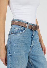 Ruskea tekstitetty vyö metallikoristeilla, jossa on suorakulmainen solki, käytetään vaaleansinisien korkeavyötäröisten farkkujen päällä. Pehmeä materiaali ja kuviollinen muotoilu.