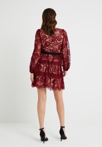 Abito in pizzo borgogna con maniche lunghe, volants a più strati, una cintura nera e orlo scallop. Indossato con sandali neri con tacco alto.