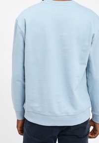 Person med en ljusblå långärmad hoodie och mörkblå jeans, sedd bakifrån mot en vit bakgrund.