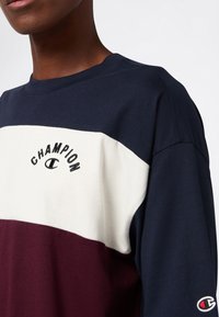 Osoba nosząca bluzę w kolorze blokowym w odcieniach granatu, bieli i burgundu z logotypami Champion na klatce piersiowej i rękawie, pokazana od szyi do połowy torsu.
