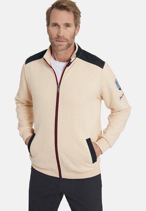 Light beige zip-up jacket with a herringbone texture, black shoulder accents, red zipper, and side pockets. Emblem on the left sleeve.
