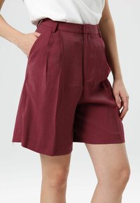Bordeauxkleurige, op maat gemaakte shorts met voorplooien, zijzakken en een gladde textuur. De tailleband zit net boven de heupen.