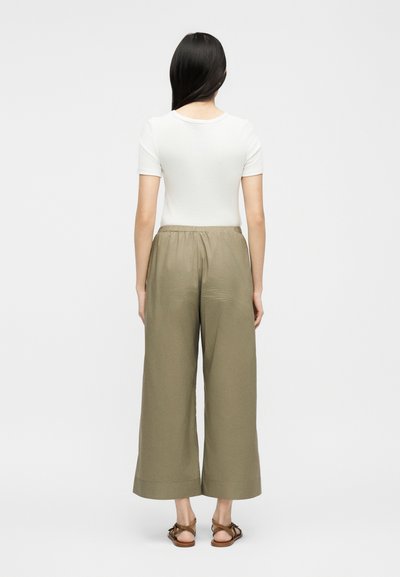 Γυναίκα με μακριά σκούρα μαλλιά, φορώντας λευκό κοντομάνικο μπλουζάκι, φαρδιά χακί παντελόνα και καφέ σανδάλια, γυρισμένη πλάτη σε μονόχρωμο φόντο.