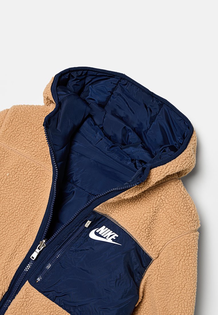 Veste à capuche en polaire marron avec doublure en nylon bleu marine, dotée d'une fermeture éclair à l'avant et du logo Nike blanc sur la partie gauche de la poitrine.