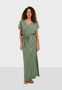 Robe maxi verte à manches courtes, décolleté en V, taille avec cordon de serrage et fente sur le côté. Tissu doux avec une texture lisse. Sandales plates visibles.