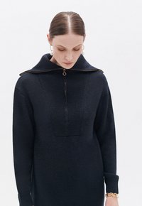 Maglione a maglia blu navy con colletto alto, dotato di una zip anteriore e pannelli testurizzati. Maniche lunghe con polsini aderenti. Dettaglio hardware dorato.