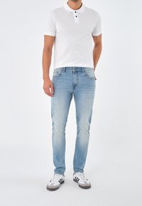 Man som bär en vit kortärmad pikétröja, ljusblå jeans och vita sneakers med svarta ränder, stående mot en enfärgad bakgrund.