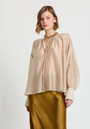 Blouse beige transparente avec un col froncé, manches longues et un détail de lien. Associée à une jupe en satin vert olive, lisse.