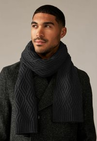 Écharpe en tricot noire avec un motif de diamant texturé enroulée autour du cou, associée à un manteau gris foncé avec un design à double boutonnage.