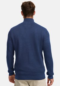 Man die een getextureerde marineblauwe gebreide trui en beige broek draagt, gezien vanaf de achterkant tegen een effen witte achtergrond.