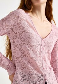 Kvinna iförd en genomskinlig rosa blommig spetsblus med lång ärm, små pärlknappar längs fram och V-ringning.