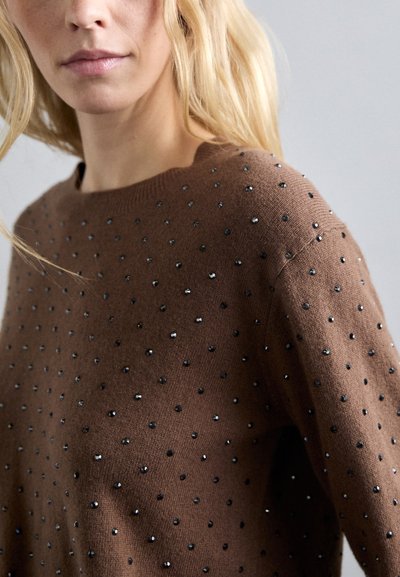 Blondynka w brązowym swetrze ozdobionym małymi, czarnymi i srebrnymi ćwiekami na jednolitym szarym tle.