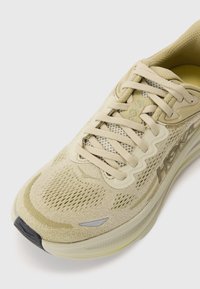 Zapatilla deportiva beige con upper de malla transpirable, diseño texturizado, cierre con cordones y un detalle de logo sutil en el costado.