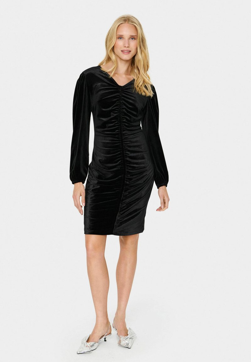 Schwarzes Samtkleid mit langen Puffärmeln und gerafftem Frontdetail, das eine taillierte Silhouette und einen knielangen Saum hat. Silberne Schuhe setzen Akzente zu dem Outfit.