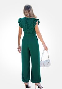 Groene jumpsuit met een kanten rug, gegolfde mouwen, wijde pijpen en een ceintuur in de taille. Gecombineerd met een kleine witte handtas en huidskleurige hakken.