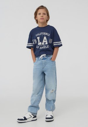 Maglietta grafica blu marina con testo bianco e maniche a strisce doppie abbinata a jeans chiari strappati e sneakers bianche con dettagli blu marina.