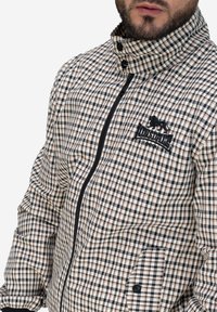 Kurtka w kratę w beżowym, czarnym i białym kolorze. Posiada wysoki kołnierz, zamek błyskawiczny, kieszenie oraz haftowane logo na klatce piersiowej.