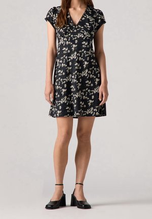 Femme portant une robe noire à manches courtes imprimée de marguerites blanches et des chaussures à talons blocs noires avec brides à la cheville, debout sur un fond neutre.