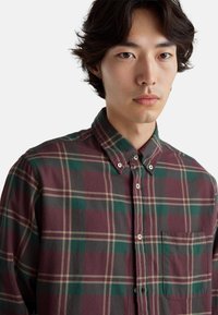 Károvaná košile s knoflíky v burgundské, zelené a šedé barvě z bavlny. Má límeček, přední kapsu a kontrastní horizontální a vertikální pruhy.