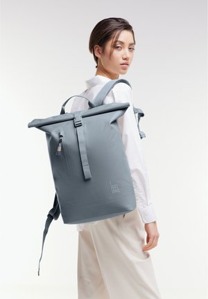 Mochila gris de tipo roll-top fabricada con material duradero, con correas ajustables, un asa en la parte superior y un logo en el frente. Textura suave.