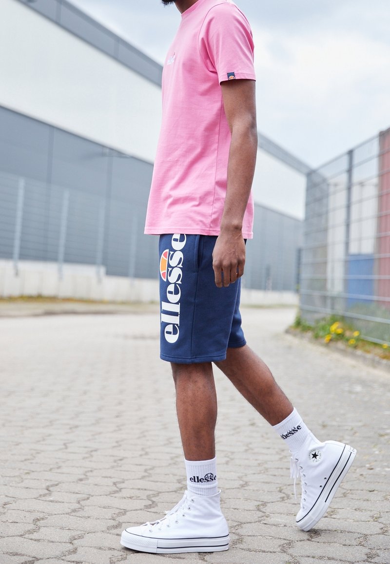 Homme portant un t-shirt rose, un short bleu marine ellesse, des baskets blanches montantes et des chaussettes blanches à la marque ellesse, debout sur un chemin extérieur pavé.