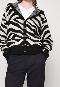 Cardigan se zebra vzorem v černé a krémové barvě, s černým žebrovaným lemem, zapínáním na knoflíky a šedou podšívkou kapuce. Nosený přes tmavé kalhoty.